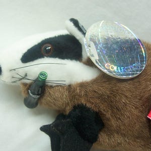 May include: A plush badger toy with a white face, black markings, and brown fur. The toy holds a miniature Coca-Cola bottle and has a tag that says "COLLECTION". A small red and white tag is attached to the toy.