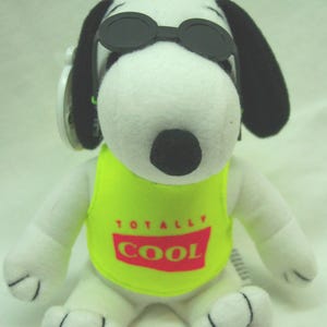 May include: A white plush dog toy with black ears and a black nose, wearing black plastic sunglasses on its head. The plush toy is dressed in a neon yellow sleeveless shirt featuring a red rectangular graphic with the text TOTALLY COOL.