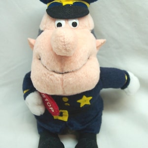 May include: A plush toy depicting a cartoon police officer. The toy is dressed in a navy blue uniform with a matching hat and a gold star. It holds a red sign that says "STOP". The toy has a large nose and a smiling face.