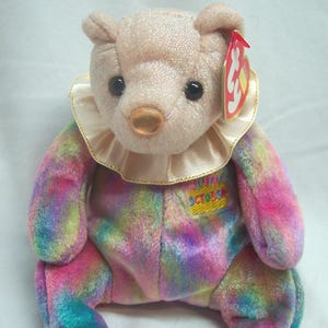 May include: A plush bear toy with a sparkly, light pink head, ears, and feet. The bear has a cream-colored ruffled collar and a multicolored body. A red tag is attached to the ear. The bear's chest has an embroidered "October" patch.
