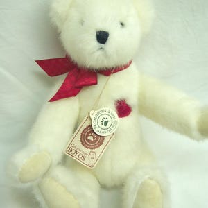 May include: A cream-colored plush teddy bear with a red ribbon around its neck and a heart-shaped patch. The bear has a black nose and a tag that reads "Boyds".