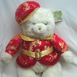 May include: A plush white teddy bear wearing a red and gold patterned outfit, including a matching hat and belt. The bear has a gold nose and is a decorative item.