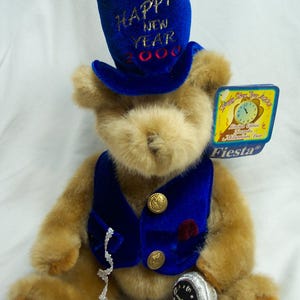 May include: A plush teddy bear wearing a blue velvet top hat and vest, with gold buttons and a small pocket watch. The hat reads "Happy New Year 2000". A tag reads "Happy New Year 2000" and "Fiesta".