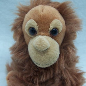 May include: A brown plush orangutan toy with large black eyes and a beige face. The toy is sitting with its legs crossed and has a soft, furry texture.