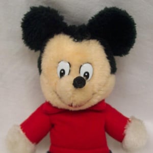May include: A plush Mickey Mouse toy wearing a red shirt and black pants. The toy has black fur, a black nose, and white fur on its hands and feet. The toy has a tag on its left leg that says 'Knickerbocker'.