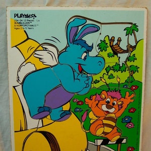 Könnte beinhalten: Ein 12-teiliges Playskool-Holzpuzzle mit Bumblelion und Hoppopot amus. Die Puzzleteile sind bunt und zeigen ein blaues Flusspferd mit Flügeln und eine gelbe Biene mit rot-weiß gestreiftem Körper. Das Puzzle ist in den USA hergestellt. Die Schachtel trägt den Hinweis "Für Kinder von 2 bis 5 Jahren".