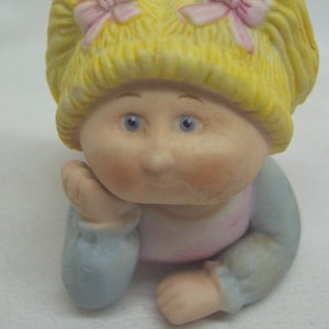 May include: A porcelain figurine of a little girl with blonde pigtails, wearing a pink dress and blue sleeves. She has blue eyes and a sweet expression.