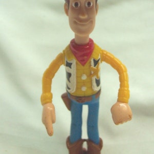 Pu&ograve; includere: Una figurina di plastica di Woody del franchise Toy Story. Woody indossa un cappello da cowboy marrone, una camicia gialla con una bandana rossa, una cintura marrone, jeans blu e stivali marroni. Ha una stella da sceriffo sul suo gilet.