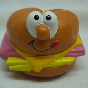 May include: A plastic toy hamburger with a smiling face, pink lettuce, yellow cheese, and a red tongue.