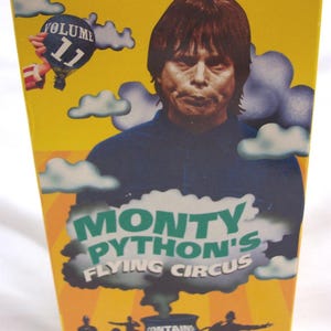 May include: A yellow VHS tape case with a picture of a man with brown hair and a blue shirt. The text "Monty Python's Flying Circus" is in green and white letters. The case says "Volume 11" and "Contains: An Exploding Penguin, On The Telly &  Bruces".