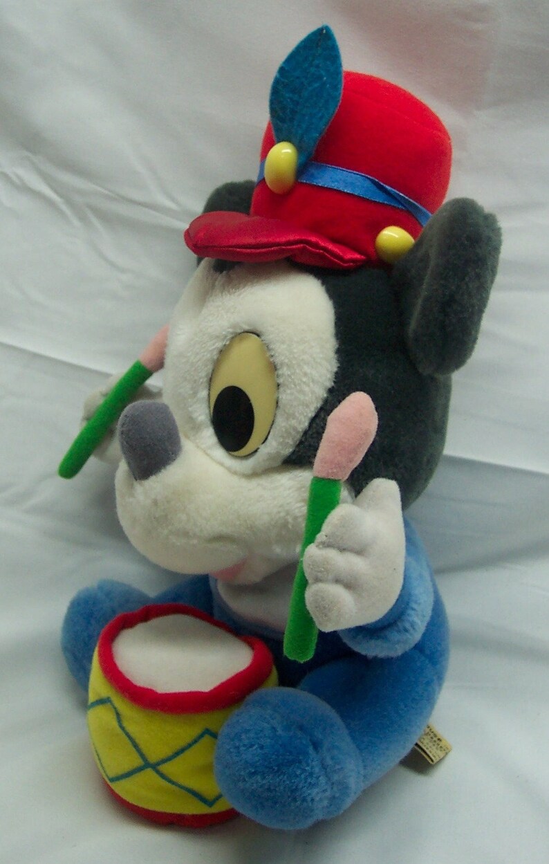 May include: A plush toy of Mickey Mouse dressed as a drummer. He is wearing a red hat with a blue band and a blue and white outfit. He is holding two drumsticks and has a small drum in front of him.