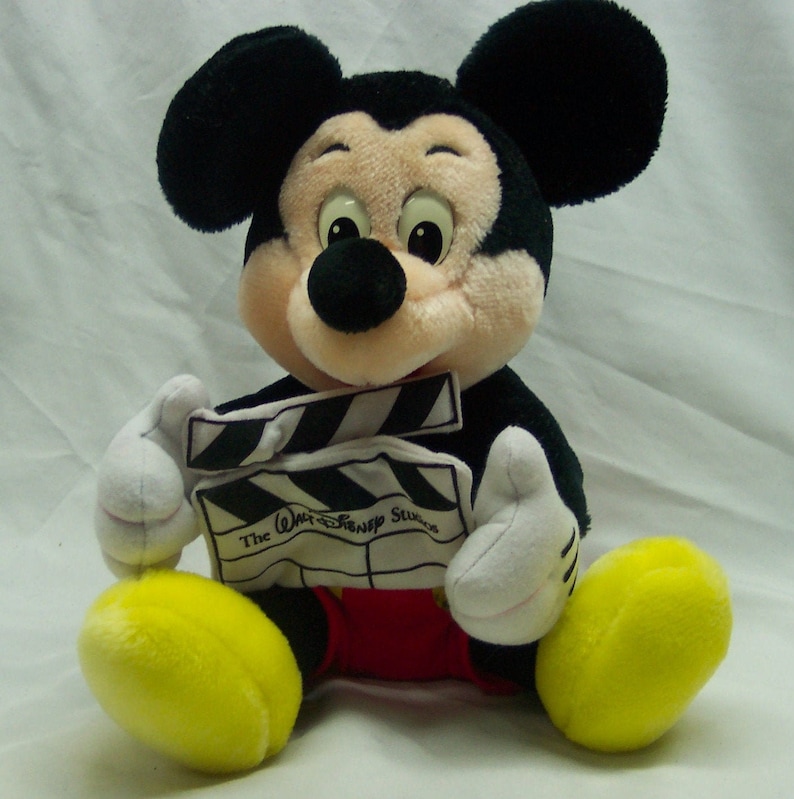 Pu&ograve; includere: Un giocattolo di peluche di Topolino che tiene una clapstick nera e bianca con il testo "The Walt Disney Studios". Topolino indossa pantaloncini rossi e scarpe gialle.