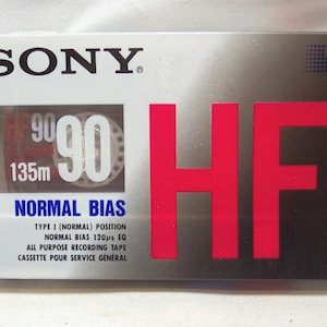 Puede incluir: Una cinta de casete de audio Sony HF 90. El embalaje es blanco y plateado con texto rojo y negro. La cinta tiene una longitud de 135 m y está etiquetada como "Normal Bias".