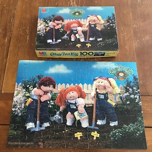 May include: A Cabbage Patch Kids 100-piece puzzle with the box on top. The puzzle and box feature illustrations of Cabbage Patch Kids dolls in a garden setting. The box has the MB logo and the words "Cabbage Patch Kids 100 Piece Puzzle."