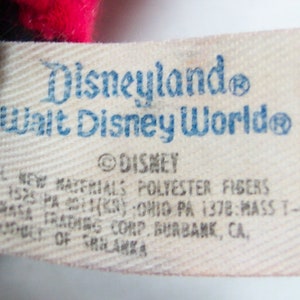 Pu&ograve; includere: Un'etichetta bianca con testo blu che recita "Disneyland Walt Disney World" e "DISNEY". L'etichetta contiene anche informazioni sui materiali utilizzati e sul produttore.