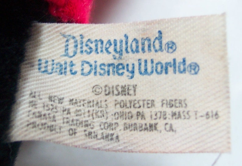 Pu&ograve; includere: Un'etichetta bianca con testo blu che recita "Disneyland Walt Disney World" e "DISNEY". L'etichetta contiene anche informazioni sui materiali utilizzati e sul produttore.