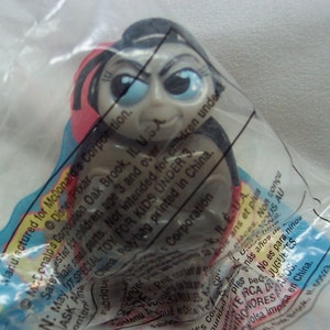 May include: A gray plastic toy figure of a character with black hair and large blue eyes. The toy is packaged in a clear plastic bag with text that reads "Manufactured for McDonald's Corporation. Disney/Pixar.  ...  Special Toy for Kids Under 3.  ...  Polybag printed in China.  ...  Corporation"