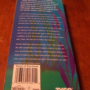 May include: A VHS tape case for the Disney movie The Little Mermaid. The case is blue and green with a picture of Ariel, the mermaid princess, on the front. The back of the case has a description of the movie and a proof-of-purchase barcode. The case also has the TYCO logo.