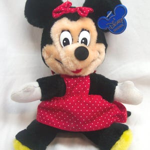 May include: A plush Minnie Mouse doll with black fur, a red polka-dot dress, and a red bow. The doll has a beige face, white gloves, and yellow soles. A blue Disney tag is attached.