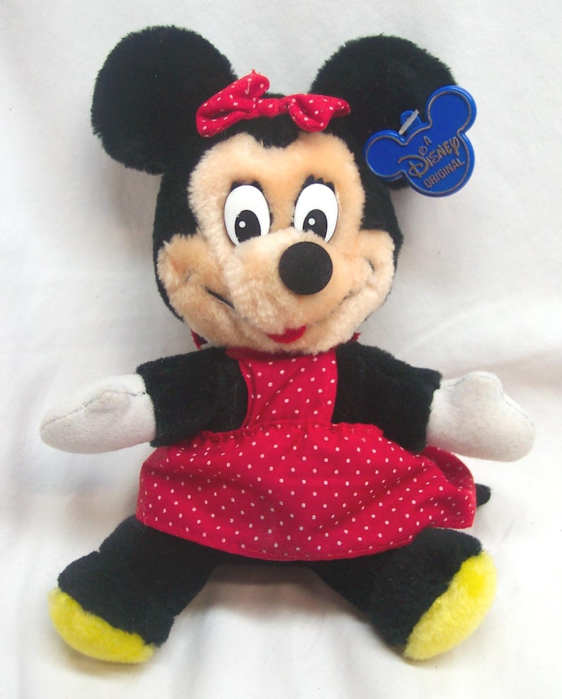 May include: A plush Minnie Mouse doll with black fur, a red polka-dot dress, and a red bow. The doll has a beige face, white gloves, and yellow soles. A blue Disney tag is attached.