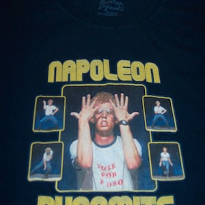 May include: Black t-shirt featuring a graphic from the movie "Napoleon Dynamite." The design includes the words "NAPOLEON DYNAMITE" in yellow block letters, with a central image of a man and smaller images around him. The man is wearing glasses and a shirt that says "VOTE FOR PEDRO."