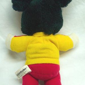 May include: A plush toy in the shape of a mouse, viewed from the back. The mouse has black ears and a black head, a yellow shirt with white sleeves, red pants, and black feet. A tag is attached to the toy.