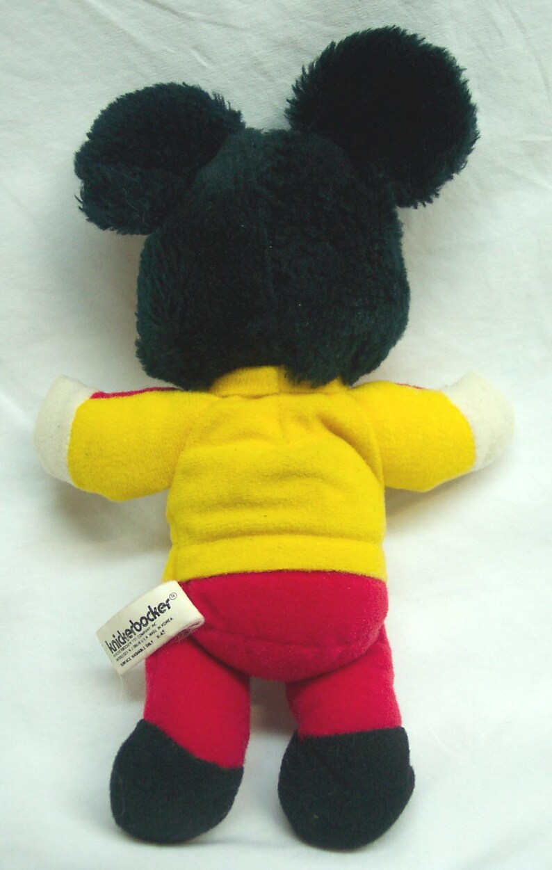 May include: A plush toy in the shape of a mouse, viewed from the back. The mouse has black ears and a black head, a yellow shirt with white sleeves, red pants, and black feet. A tag is attached to the toy.