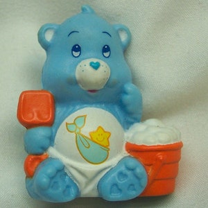 May include: A blue Care Bear with a light blue whale and a yellow star on its belly. The bear is holding a red and orange bucket with white foam inside. The bear is wearing a diaper and has a red and orange shovel in its left hand.