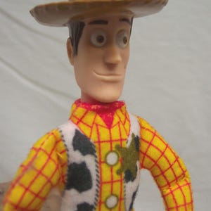Puede incluir: Figura de acci&oacute;n de Woody, con sombrero de vaquero marr&oacute;n, camisa a cuadros amarilla con detalles rojos, chaleco blanco con manchas de vaca negras y pantalones azules. La figura tiene una expresi&oacute;n sonriente.