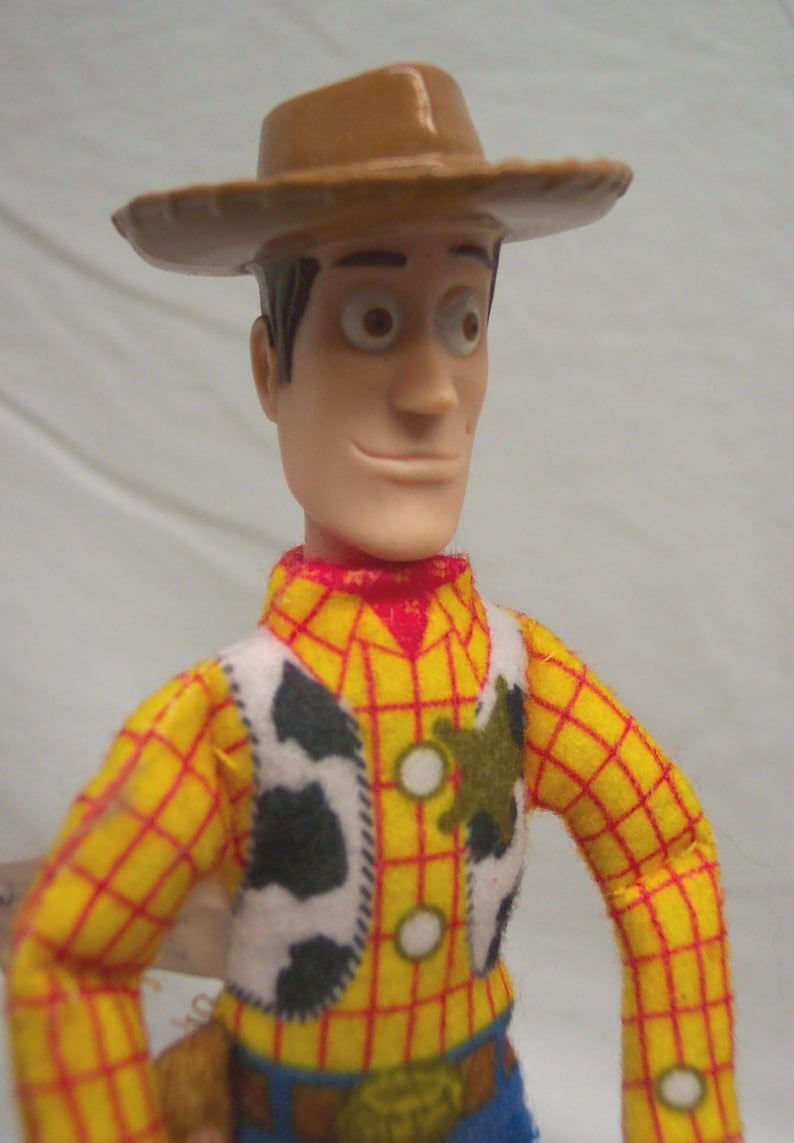 Puede incluir: Figura de acci&oacute;n de Woody, con sombrero de vaquero marr&oacute;n, camisa a cuadros amarilla con detalles rojos, chaleco blanco con manchas de vaca negras y pantalones azules. La figura tiene una expresi&oacute;n sonriente.