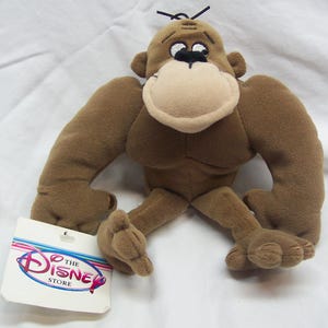 May include: Brown plush gorilla toy with a large, round nose and big eyes. The toy is sitting with its legs outstretched. The toy has a tag attached to its leg that reads "The Disney Store".