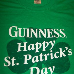 May include: Green t-shirt with the Guinness logo and the words "Happy St. Patrick's Day" in white. A large green shamrock shape is behind the text. The shirt has a crew neck.