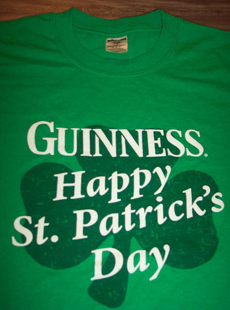 May include: Green t-shirt with the Guinness logo and the words "Happy St. Patrick's Day" in white. A large green shamrock shape is behind the text. The shirt has a crew neck.