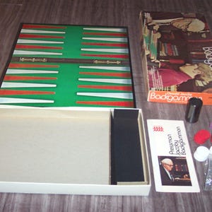 Puede incluir: Un juego de backgammon vintage con un tablero de juego verde y rojo, una caja blanca con un interior negro y una caja de fichas. La caja del juego tiene el texto "Pressman Family Games Backgammon" impreso.