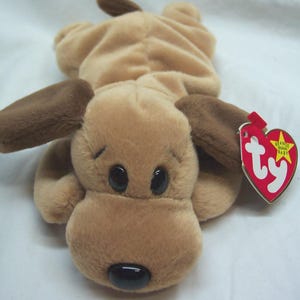 May include: A brown and tan plush dog toy with a brown nose and black eyes. The toy has a Ty Beanie Baby tag attached to it.