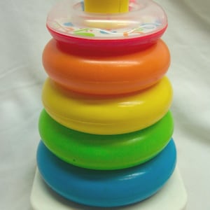 May include: A classic Fisher-Price stacking toy with a white base and a series of colourful rings. The rings are in shades of blue, green, yellow, orange, and a clear top ring with a red rim. A yellow peg tops the stack. The Fisher-Price logo is visible.