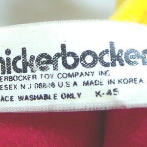 May include: Close-up of a white tag with the word "knickerbocker" in black letters. The tag also includes the text "KNICKERBOCKER TOY COMPANY INC MIDDLESEX N.J. 08846 U.S.A. MADE IN KOREA SURFACE WASHABLE ONLY K-45". The tag is on a red and yellow background.