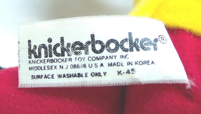 May include: Close-up of a white tag with the word "knickerbocker" in black letters. The tag also includes the text "KNICKERBOCKER TOY COMPANY INC MIDDLESEX N.J. 08846 U.S.A. MADE IN KOREA SURFACE WASHABLE ONLY K-45". The tag is on a red and yellow background.