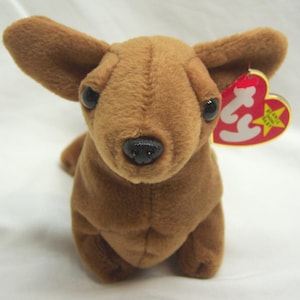 May include: A brown plush Chihuahua dog with large ears and black eyes. The dog has a black nose and a red heart-shaped tag with white text and a yellow star. The plush toy is soft and cuddly.