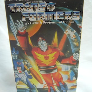 May include: A vintage VHS tape box for "The Original Transformers: Volume 3: Programmed For Evil." The box features colorful illustrations of Transformers characters in action, with the title text prominently displayed. The Kid Rhino Home Video logo is visible.