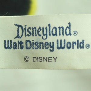 May include: A white tag with the words "Disneyland" and "Walt Disney World" printed in blue. The tag also includes the Disney copyright symbol and the word "DISNEY". The tag is attached to a dark-colored plush toy.