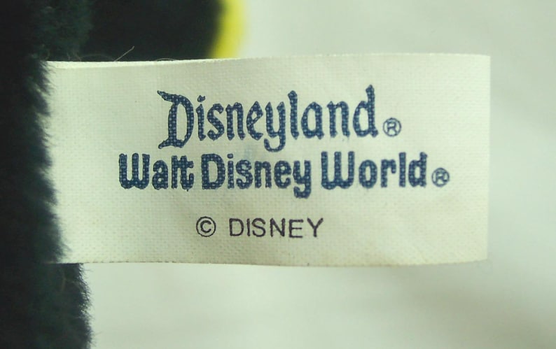 May include: A white tag with the words "Disneyland" and "Walt Disney World" printed in blue. The tag also includes the Disney copyright symbol and the word "DISNEY". The tag is attached to a dark-colored plush toy.