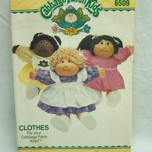 May include: A Butterick sewing pattern envelope for Cabbage Patch Kids clothes, number 6509. The front features three dolls wearing different outfits: a yellow dress with a white apron, a purple floral dress with a white apron, and a pink polka dot dress.