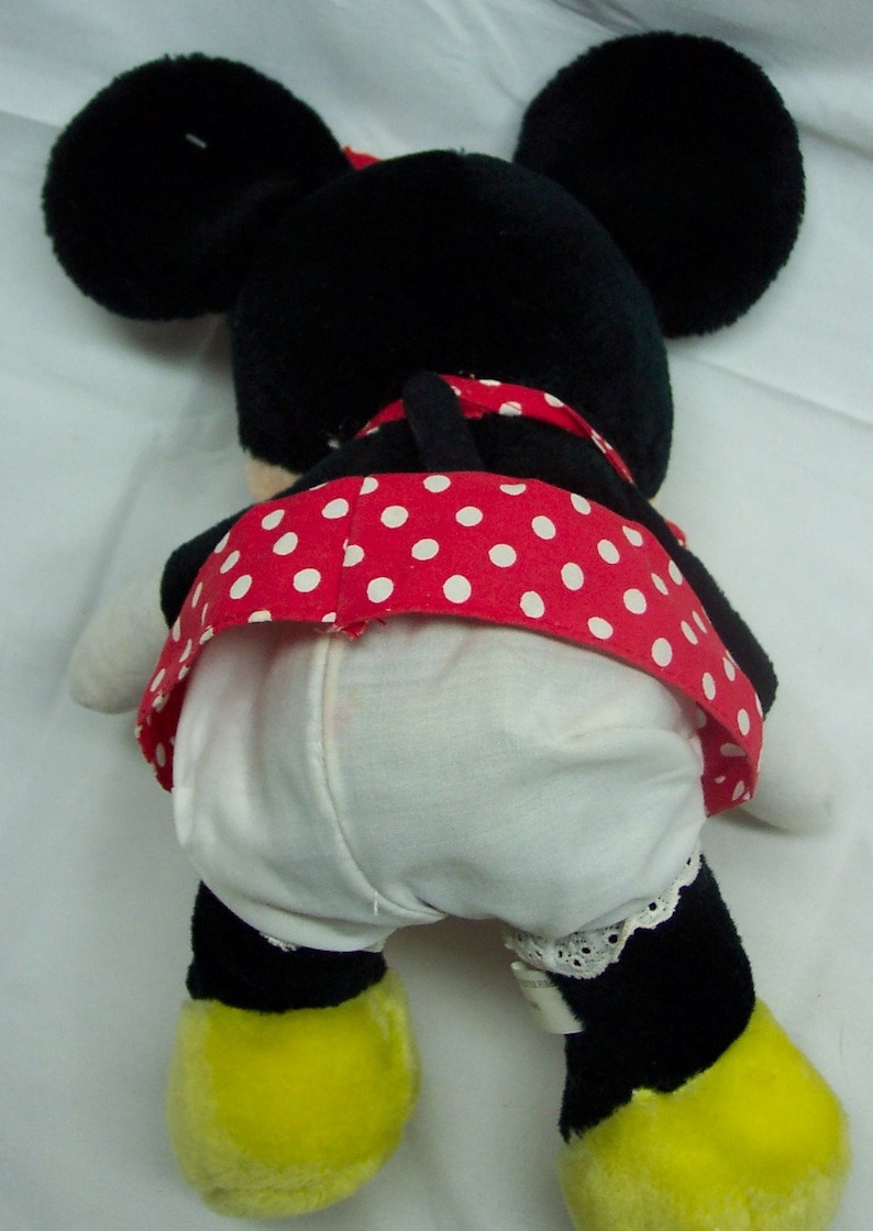 May include: A black and white plush toy of a cartoon mouse wearing a red and white polka dot dress. The toy has yellow feet and is sitting on a white surface.