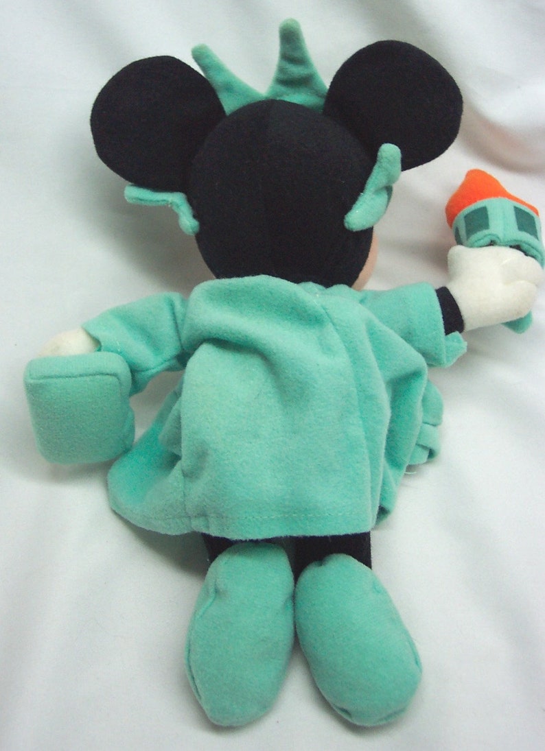Peut inclure: Une peluche de Mickey Mouse habill&eacute;e en statue de la Libert&eacute;. La peluche porte une robe verte avec une couronne et tient un flambeau miniature.