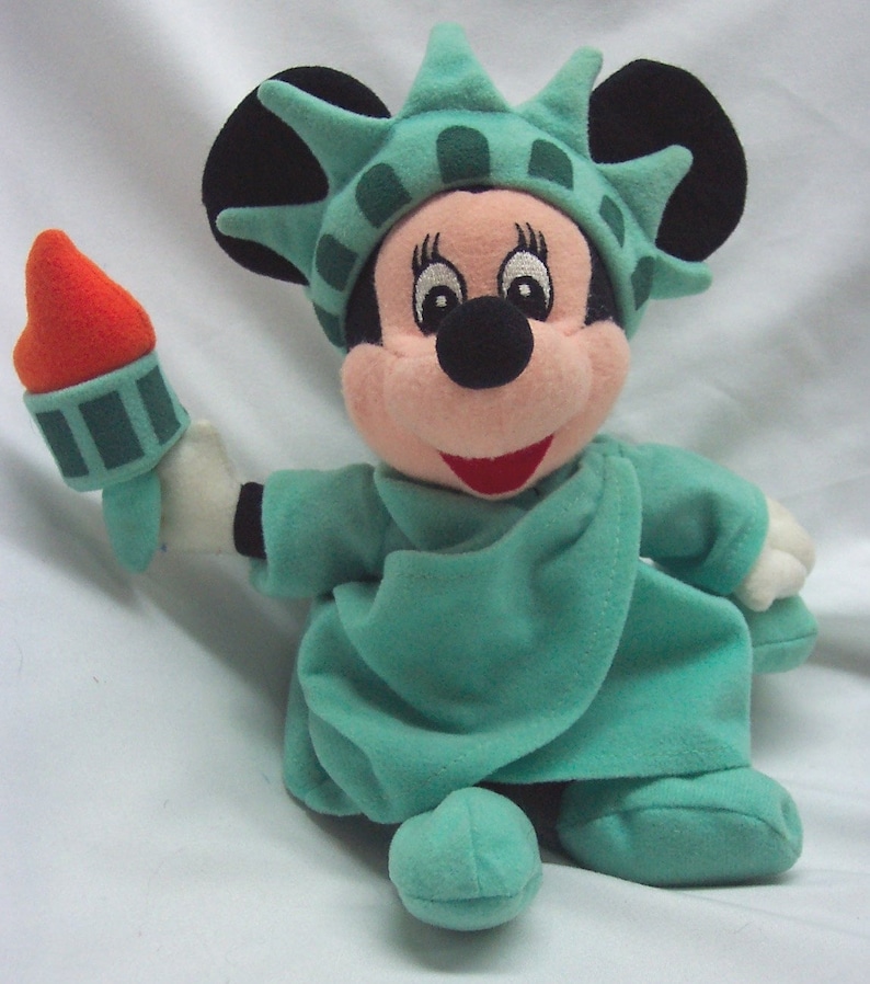 Peut inclure: Un jouet en peluche de Mickey Mouse habill&eacute; en statue de la Libert&eacute;. Le jouet tient une torche dans sa main droite. Le jouet porte une robe verte et une couronne avec des pointes. Le jouet a un nez rouge et des yeux noirs.