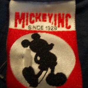 May include: A white rectangular clothing tag with red accents featuring the text MICKEY INC and SINCE 1928 in black lettering. The center of the tag displays a black silhouette of Mickey Mouse inside a white circle against a red background.