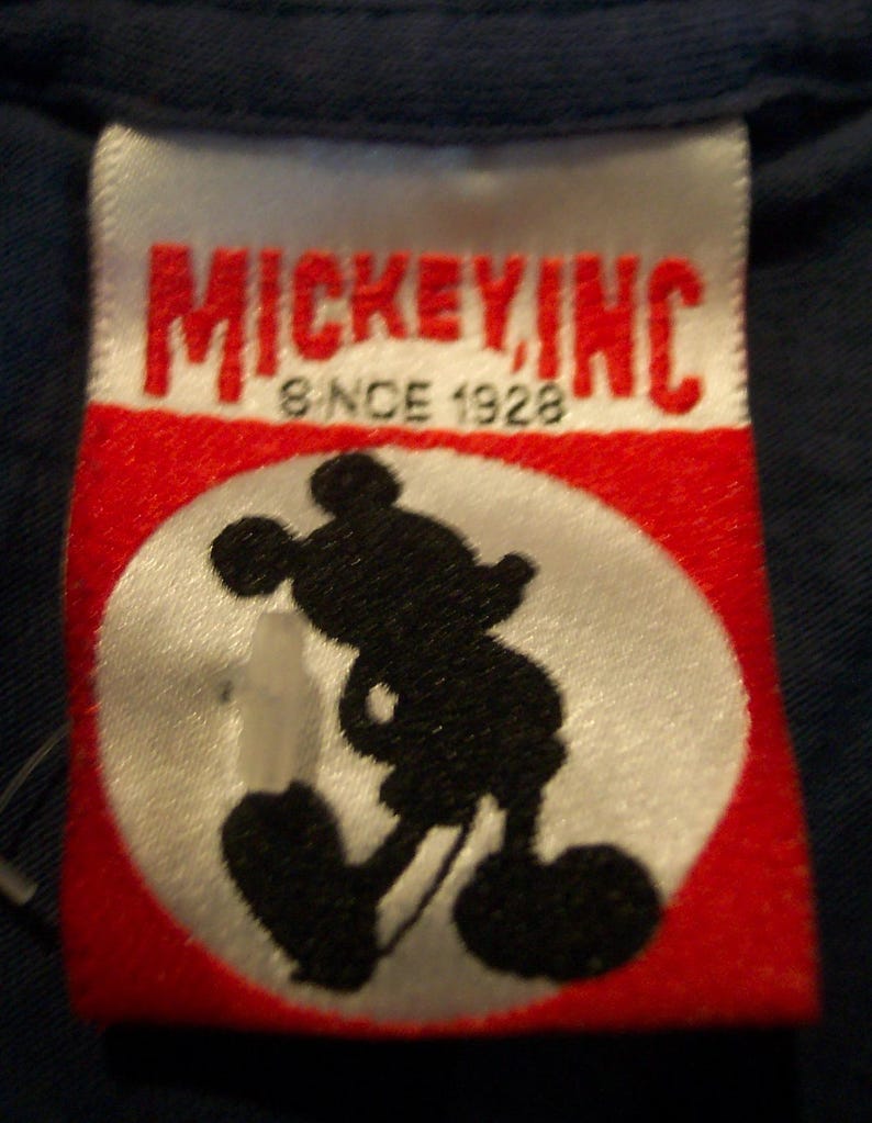 May include: A white rectangular clothing tag with red accents featuring the text MICKEY INC and SINCE 1928 in black lettering. The center of the tag displays a black silhouette of Mickey Mouse inside a white circle against a red background.