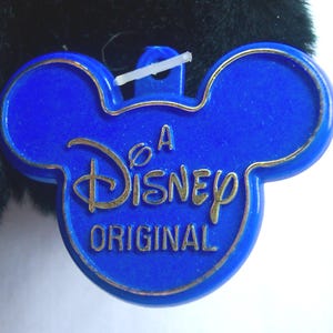 May include: A blue Disney Original tag in the shape of Mickey Mouse's head. The tag has a gold border and gold lettering that reads "A Disney Original". The tag is attached to a plush toy.