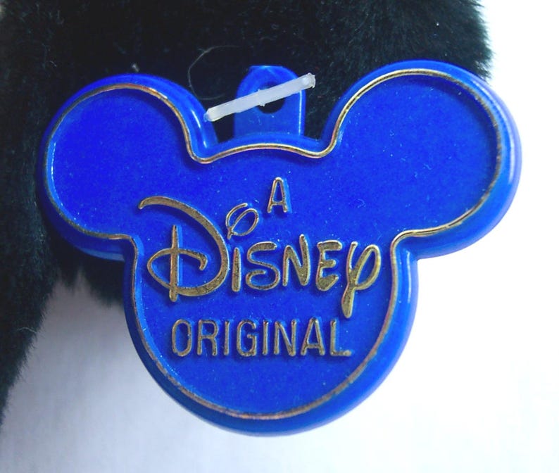 May include: A blue Disney Original tag in the shape of Mickey Mouse's head. The tag has a gold border and gold lettering that reads "A Disney Original". The tag is attached to a plush toy.